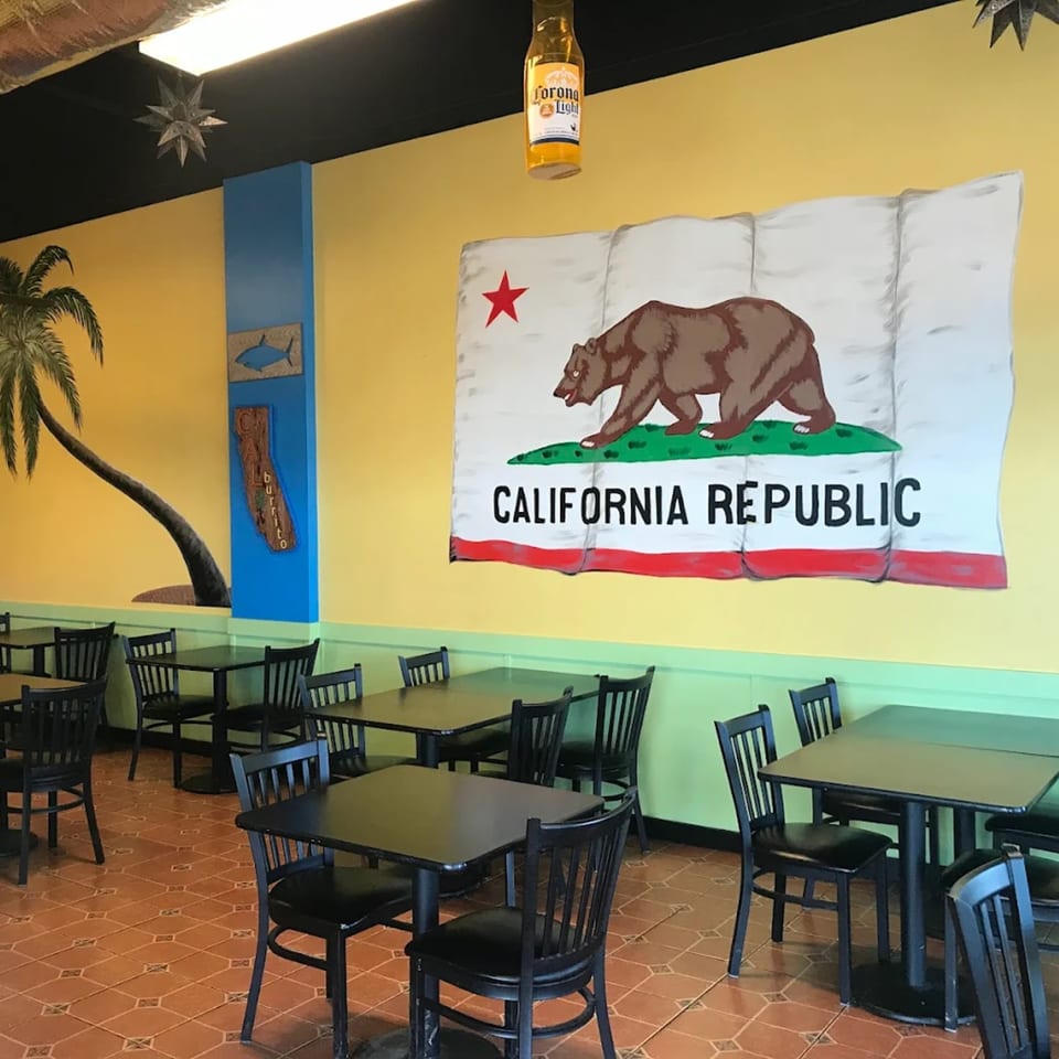 Interior of Cali Burrito Mexican Grill with a large California Republic flag mural and black tables.