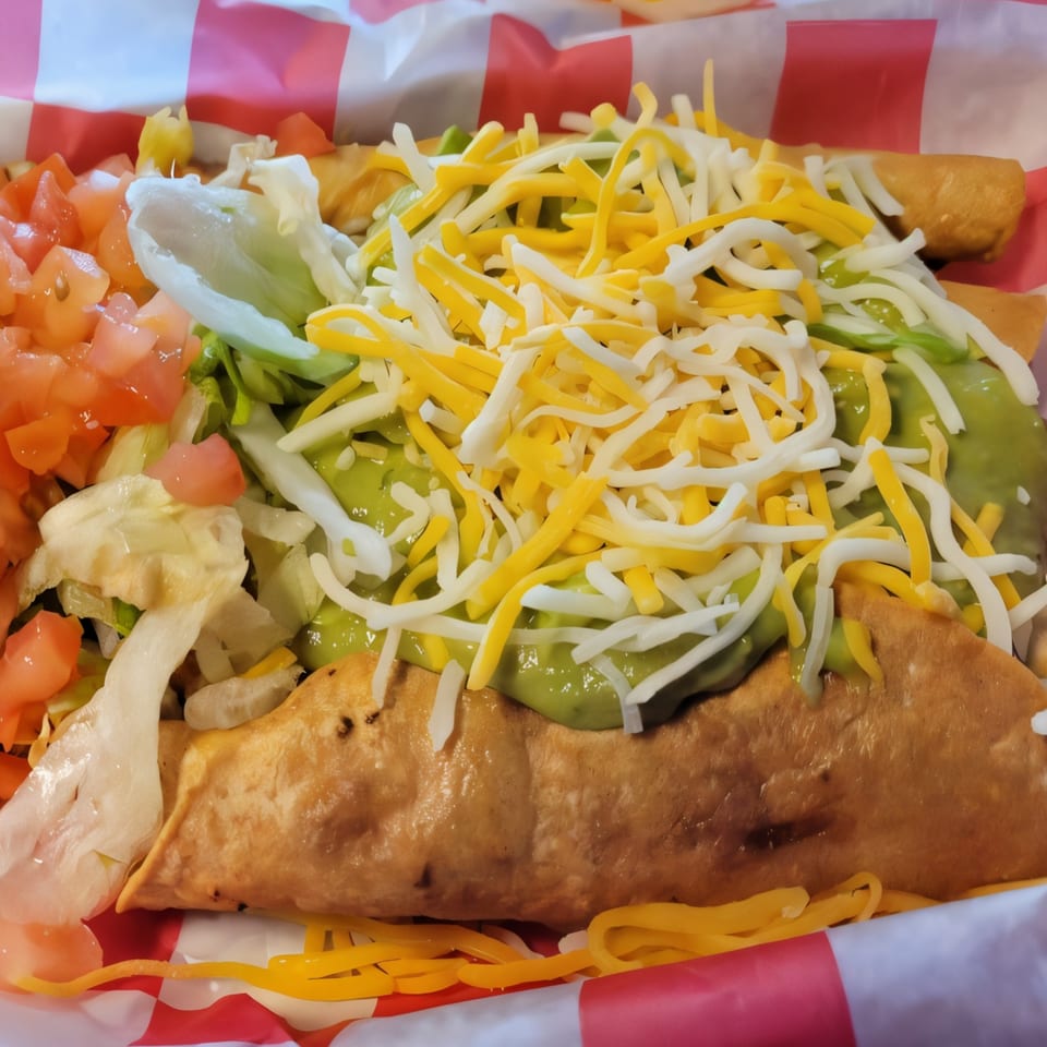 Crispy taquitos topped with guacamole, shredded cheese, lettuce, and diced tomatoes in a red and white basket.